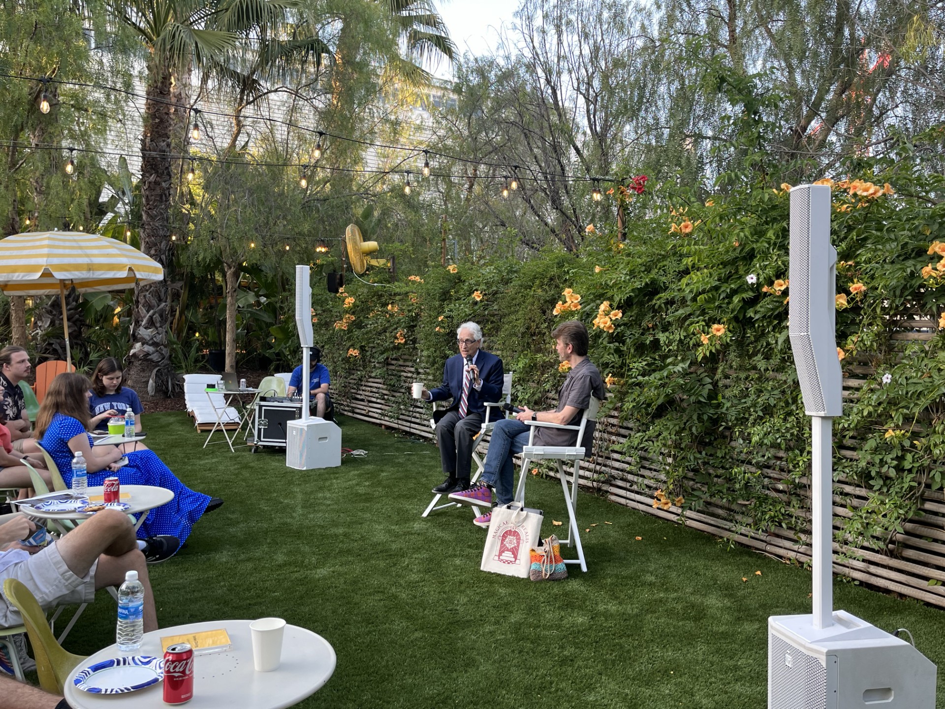 Two people sit in directors' chairs and talk in front a green plant wall