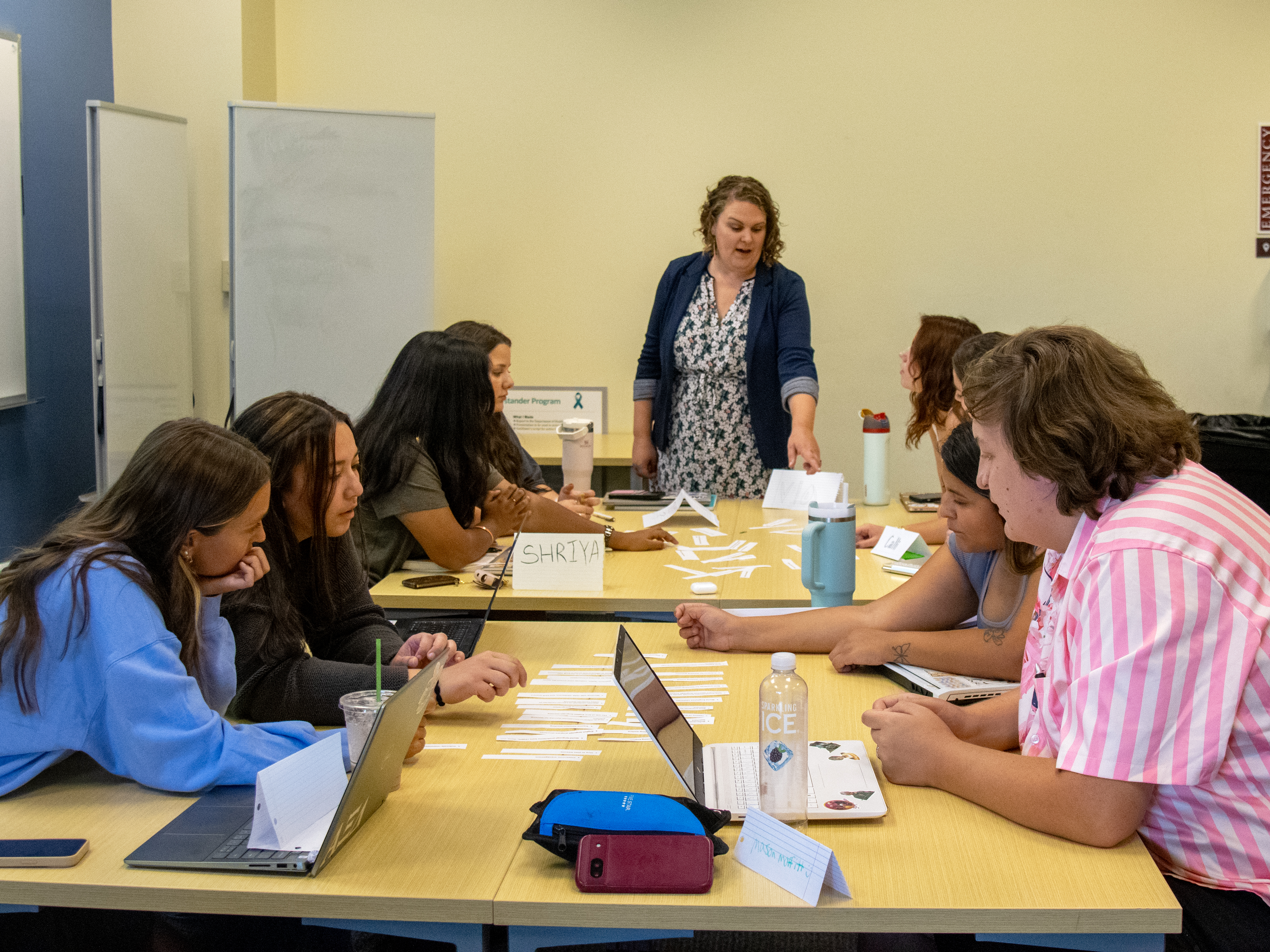 8 students around tables and a woman in a dress and blue sweater instructing them