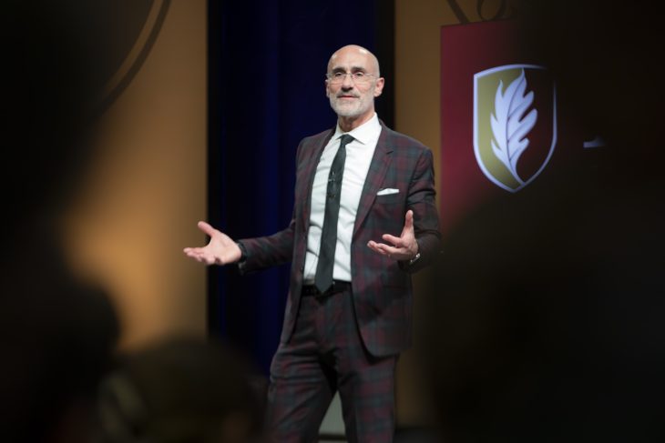 Arthur C. Brooks speaks with arms out in front of Elon sign