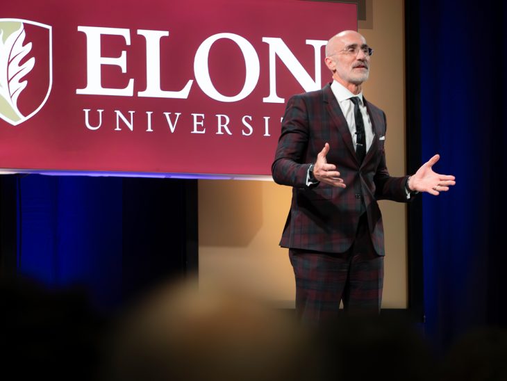 Arthur C. Brooks speaks with arms out in front of Elon sign