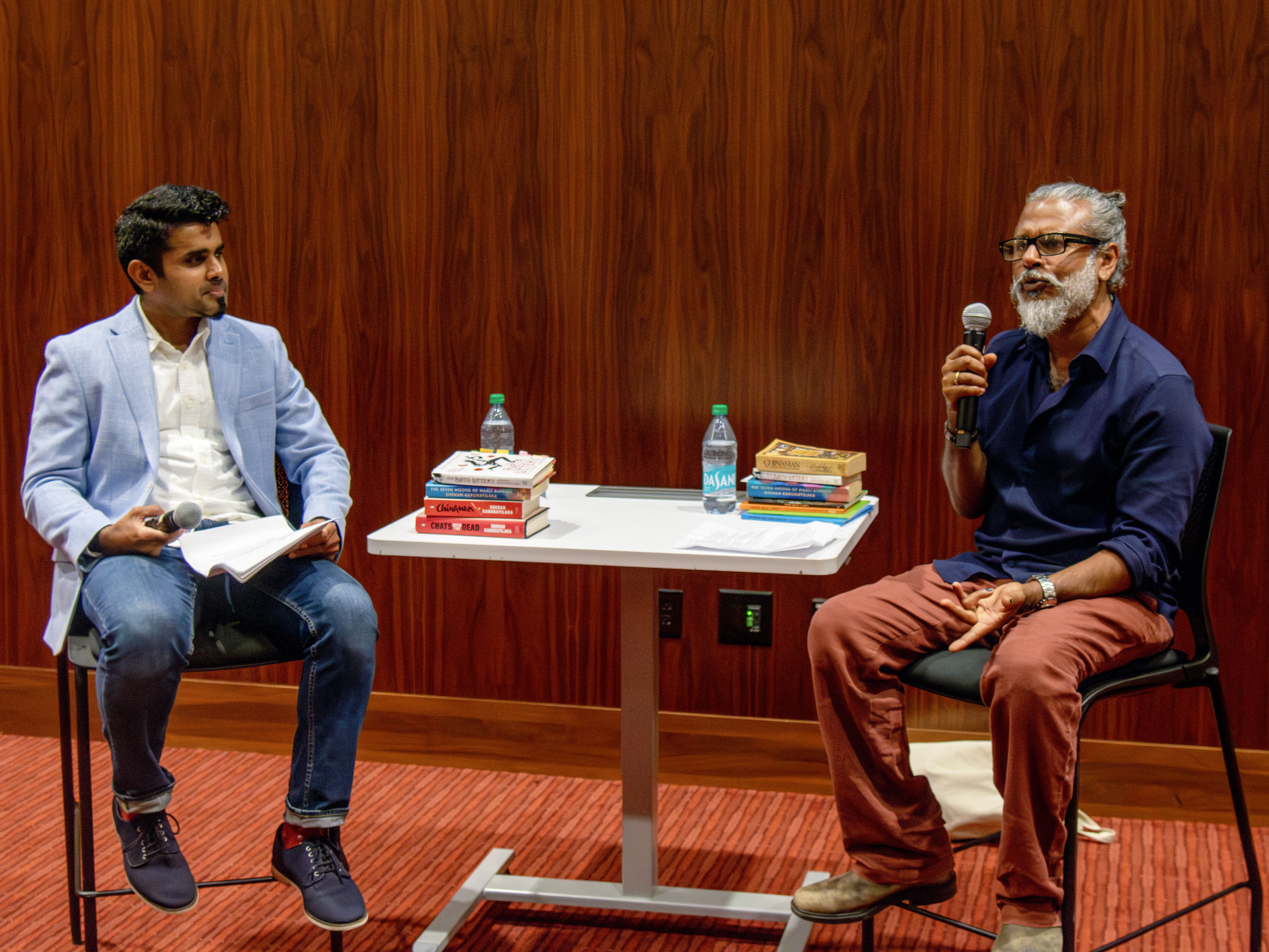 Two men with microphones at a table stacked with books