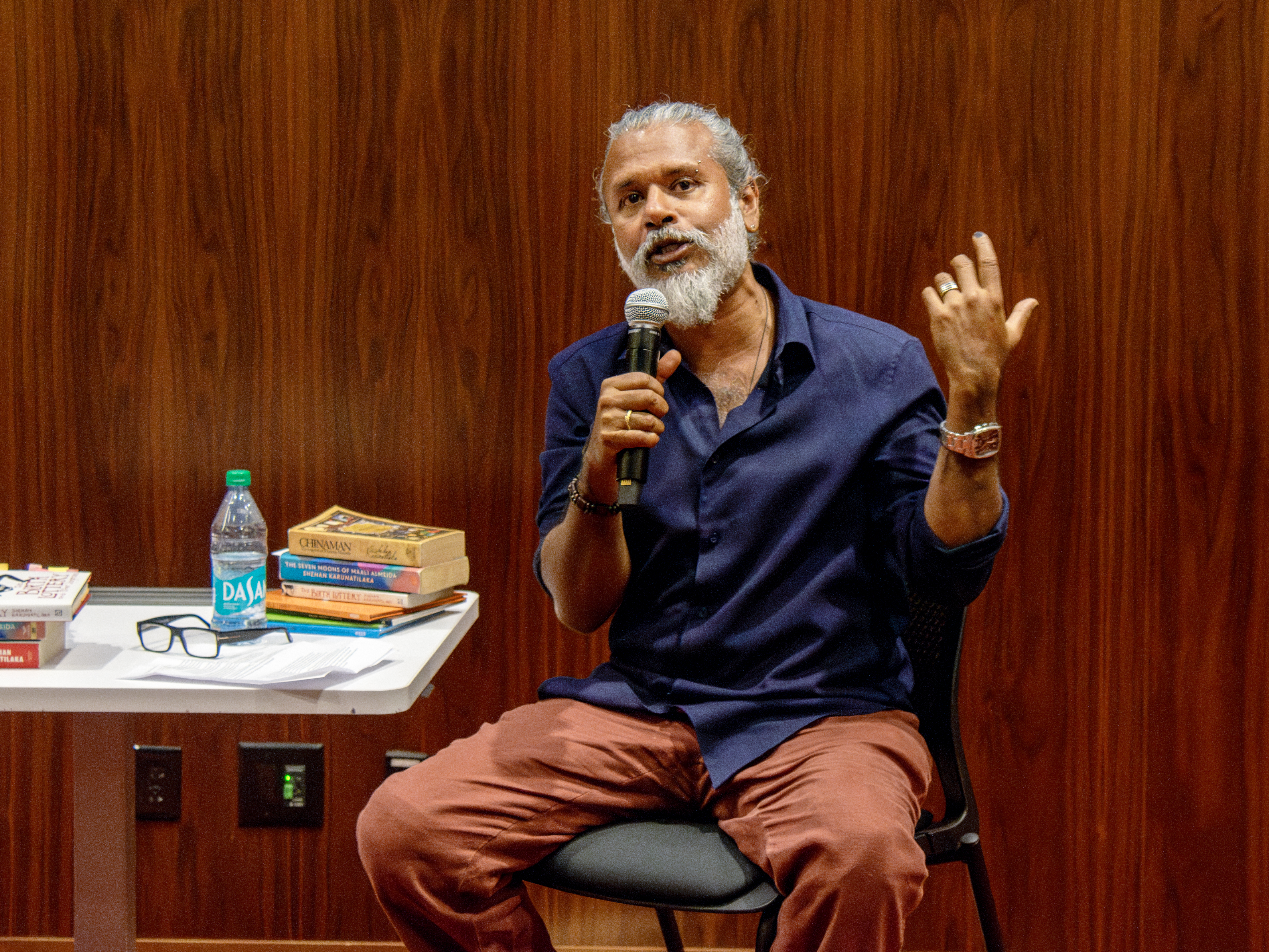 Shehan Karunatilaka speaks into a microphone while seated at a table stacked with books