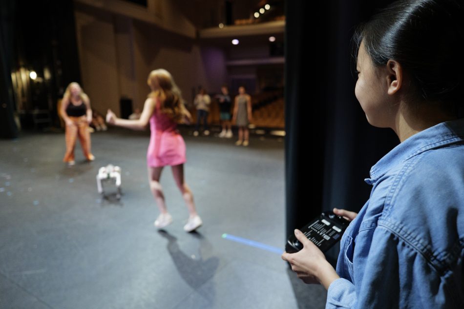 A student uses a remote control and watches a robot dog onstage with two actors