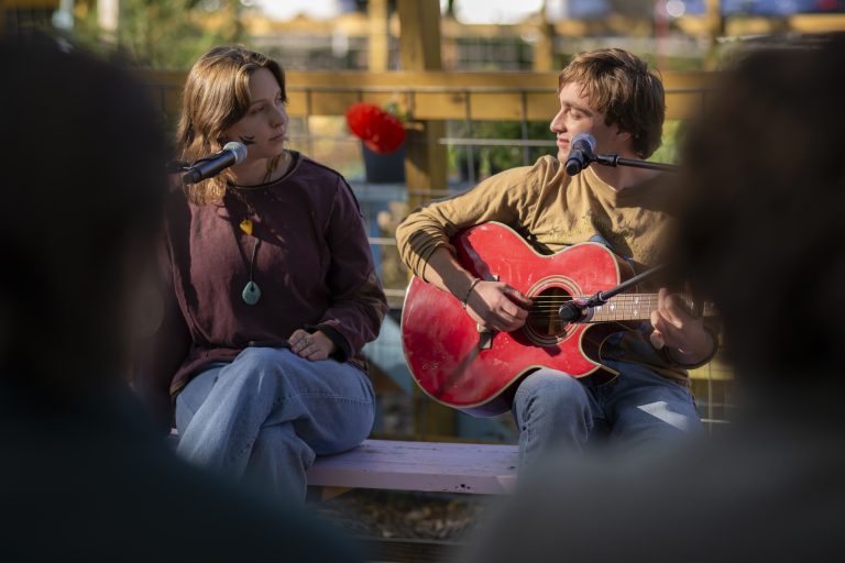 Person plays a guitar while person next to him looks on