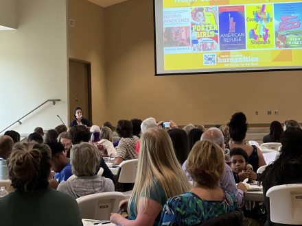 A crowd of people at round tables listen to an author speak at a podium in a large fellowship hall