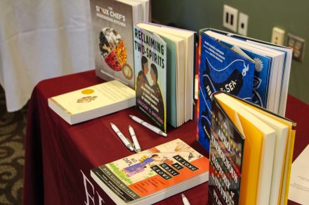 A display table covered with a red cloth holds a selection of books focusing on Indigenous and cultural themes. Titles visible include "The Sioux Chef's Indigenous Kitchen," "Reclaiming Two-Spirits," "As Long as Grass Grows," "An Afro-Indigenous History of the United States," and others. Several pens are scattered around the table, suggesting the display may be for promotional or educational purposes.