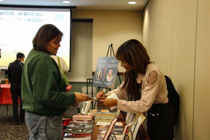 Two women are interacting at a table displaying books and informational materials for Native American Heritage Month. One woman, wearing a green sweater, examines a pamphlet handed to her by the other woman, who is wearing a light-colored sweater. A large screen in the background displays “Native American Heritage Month.” Other attendees and materials are visible in the room.