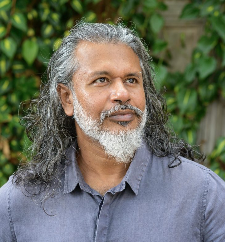 Shehan Karunatilaka in a blue shirt next to an ivy-covered wall