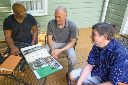 Three men sit around a table looking at a scrapbook