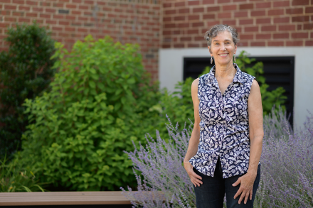 A woman poses for a portrait outdoors in front of a brick building and some green bushes