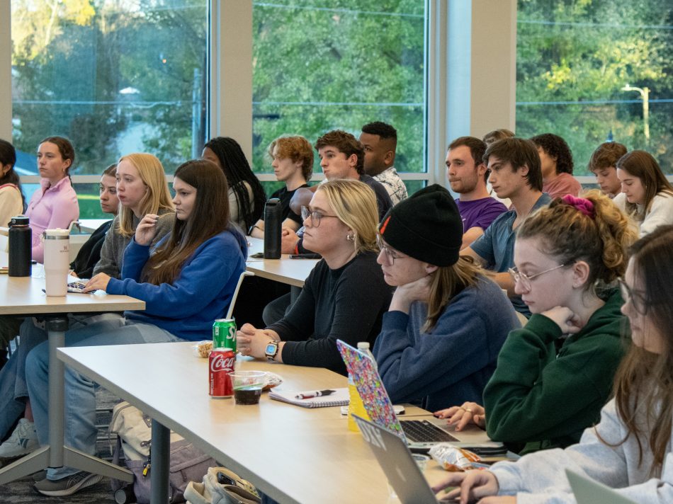 A large audience of students is seated in rows, focused on a speaker or presentation. Some individuals are taking notes on laptops, while others hold drinks and snacks.