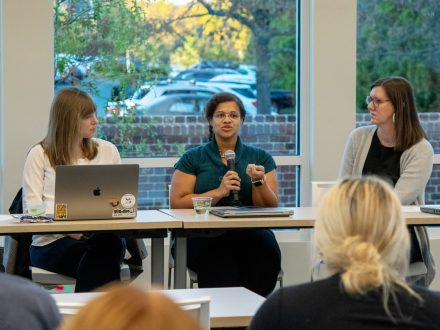 Three women panelists sit at a table in front of an audience. The woman in the center is speaking into a microphone, while the others listen. A laptop with stickers is open on the table.