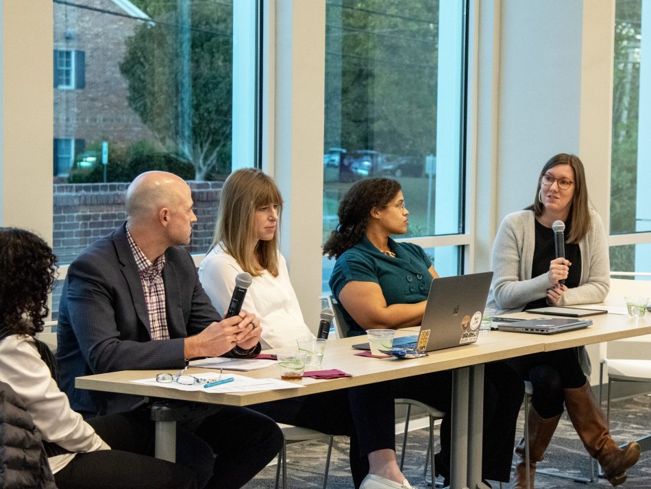 five people, four women and one man, sit at a long table with microphones. One is using a laptop.