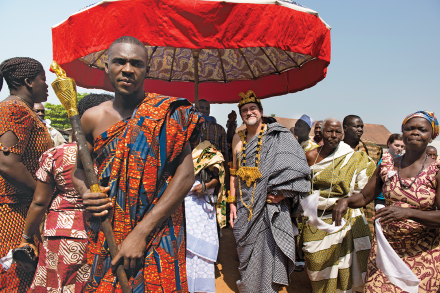 A group of people in traditional Ghanaian clothing stand outside beneath a red umbrella