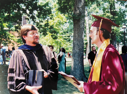 A male university graduate in commencement robes talks with a male professor wearing academic regalia in front of an oak tree 