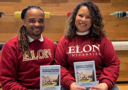 Stephanie Hernandez Rivera, assistant professor in the Master of Higher Education Program and the Dr. Jo Watts Williams School of Education Emerging Professor (right), and Jonathan A. McElderry, dean of Student Inclusive Excellence and assistant professor (left), seated on tiered wooden seating. Both wear red "Elon University" sweatshirts and hold copies of their book, Developing an Intersectional Consciousness and Praxis: Moving Toward Antiracist Efforts in Higher Education.