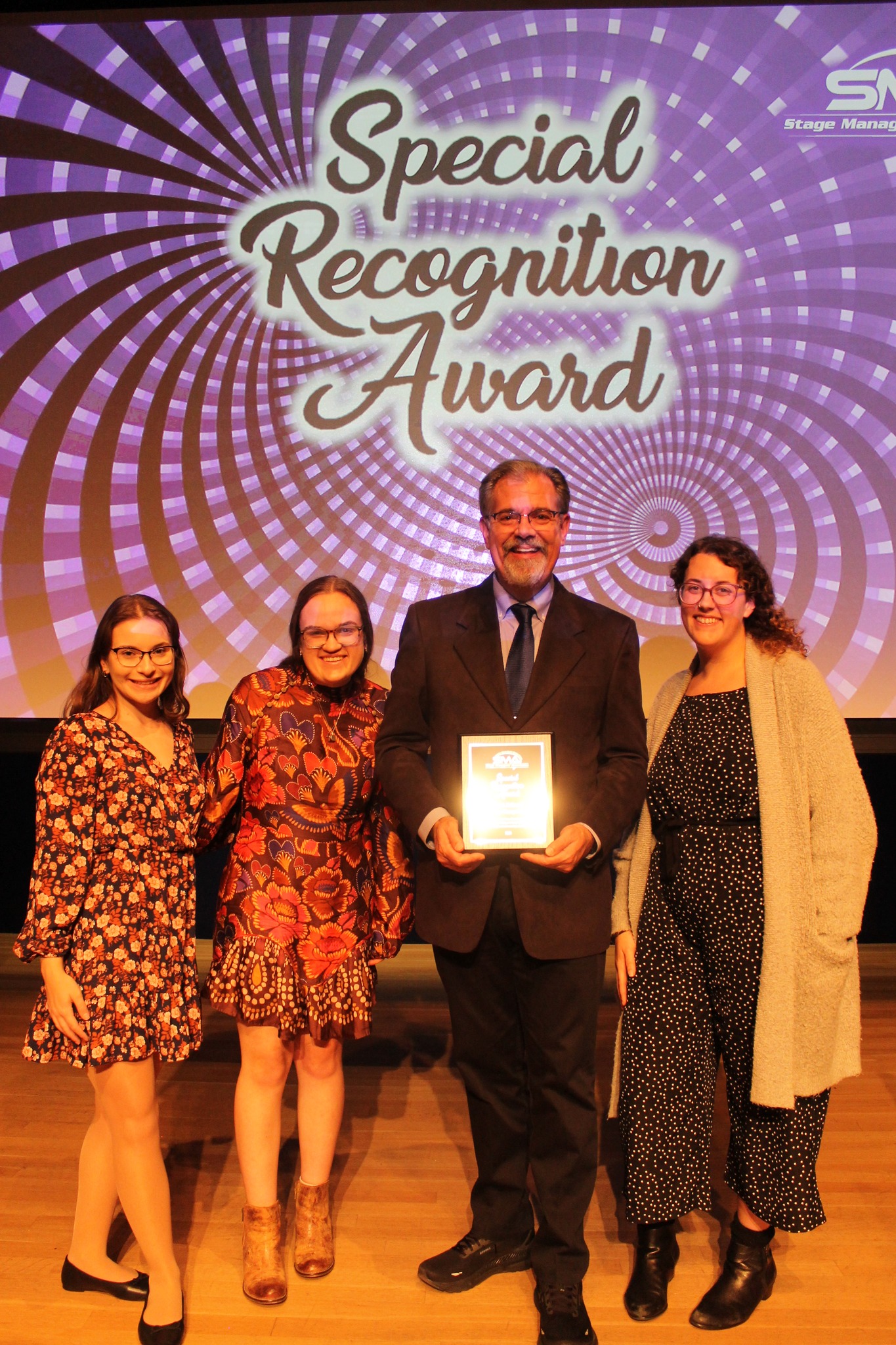 Three women stand with a man in a suit holding a plaque in front of a purple and black screen that says Special Recognition Award