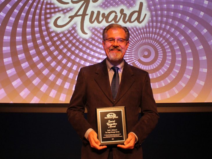 A man in a gray suit and blue tie holds a plaque in front of a projected screen of swirling purple and black that says Special Recognition Award