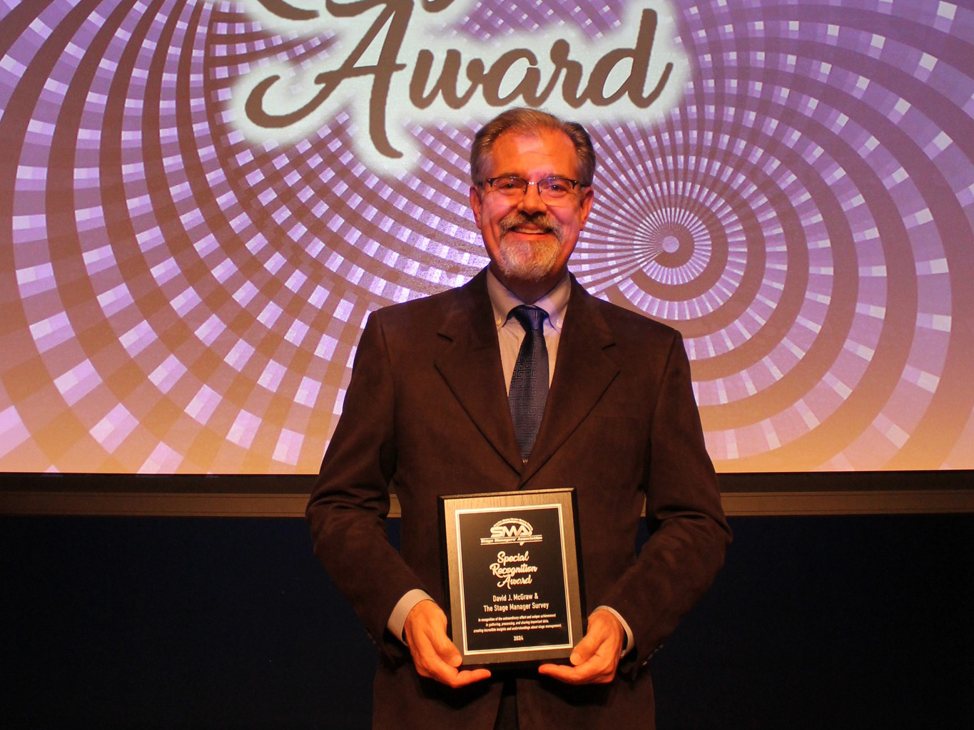 A man in a gray suit and blue tie holds a plaque in front of a projected screen of swirling purple and black that says Special Recognition Award