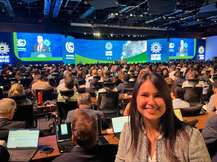 A woman with brown hair turned and smiling at the camera as a crowded conference hall seated at long tables watches U.S. President Joseph Biden on screens.