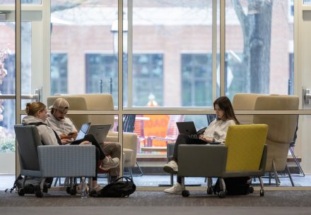 Three students sit in a lounge area, working on laptops and devices. Two sit together in gray armchairs, while the third sits alone in a yellow-backed chair. A large window behind them reveals a campus courtyard with outdoor furniture and bare trees. The atmosphere suggests focused study or collaboration.