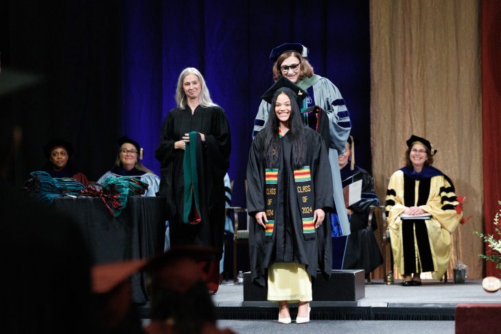 Two women stand behind another. All are wearing regalia. One woman in regalia places a hood on another one.