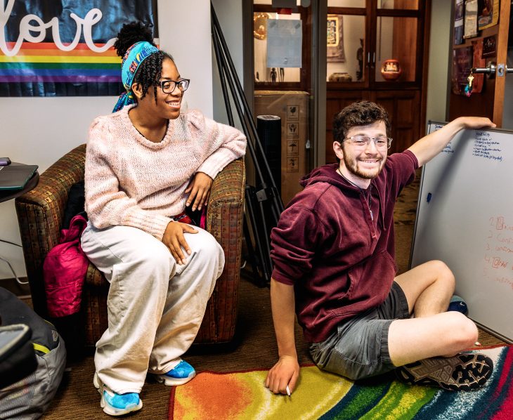 Morgan Williams sits in a chair and looks at whiteboard. Ryan Gibbons holds a whiteboard while sitting on the floor.