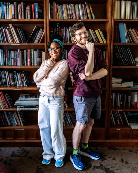 Two people stand side by side with their hands on their chins. A bookshelf is behind them