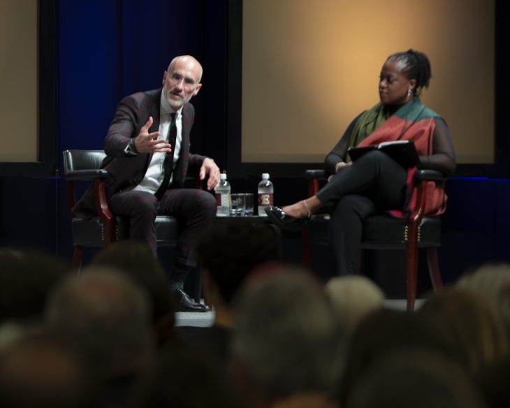 Two people sit in chairs. The main on the left is speaking to an audience with his hand outstretched. The woman on the right is looking at him.