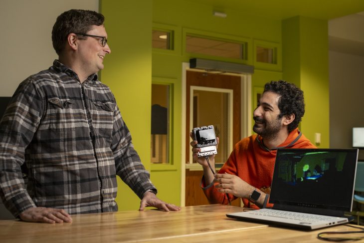 Ryan Mattfield stands with hands on a table. Rony Dahdal sits while holding LiDAR technology. A computer is sitting next to Dahdal. 