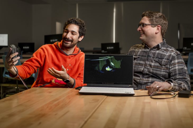 Rony Dahdal sits next to Ryan Mattfield at a table. A laptop sits between them. Dahdal is showing a square device to Dahdal. 