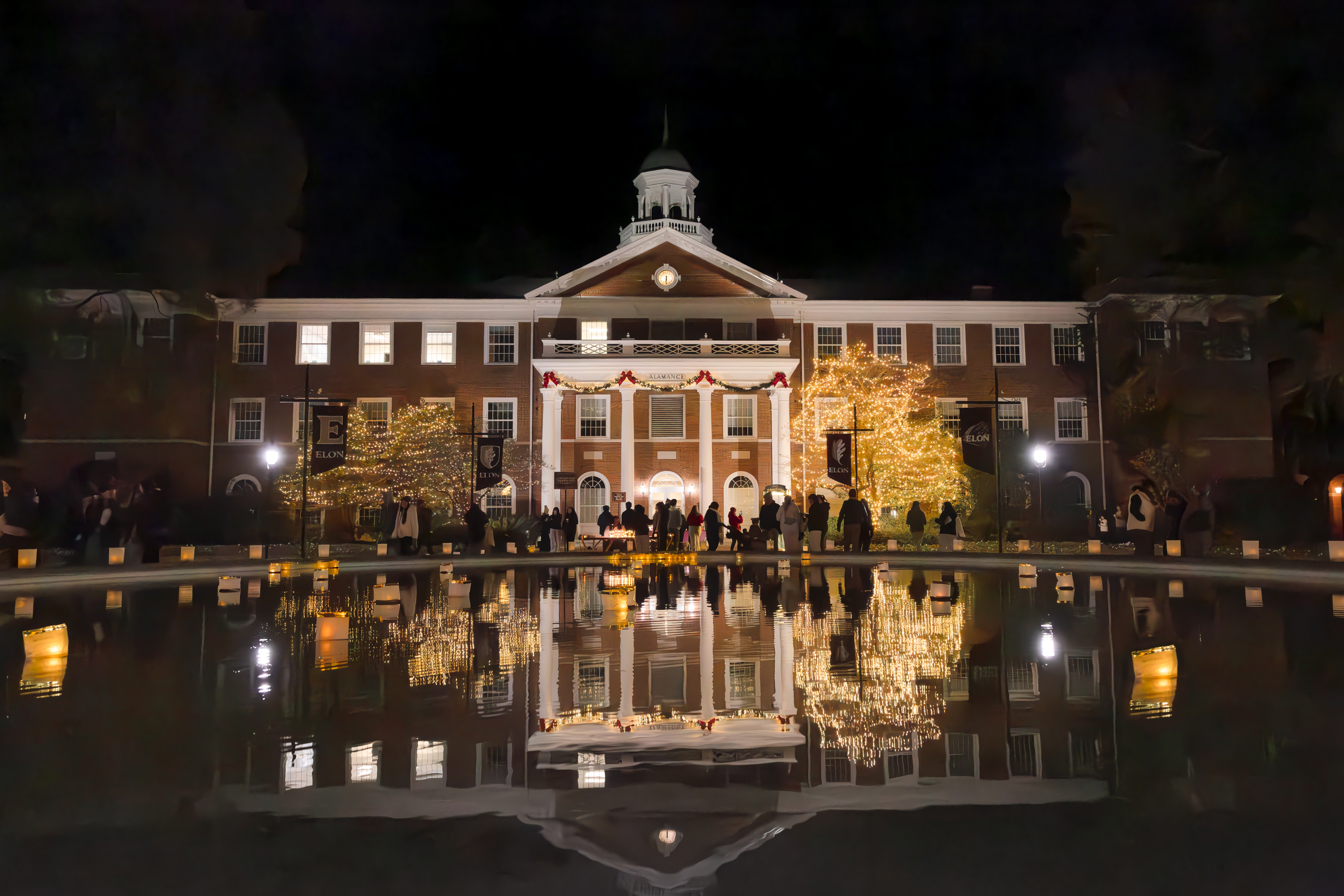 Front of Alamance Building at Elon University illuminated with festive lights and decorations, reflected in a pool of water. Crowds gather to celebrate the Festival of Lights and Luminaires, with glowing lanterns lining the walkway.