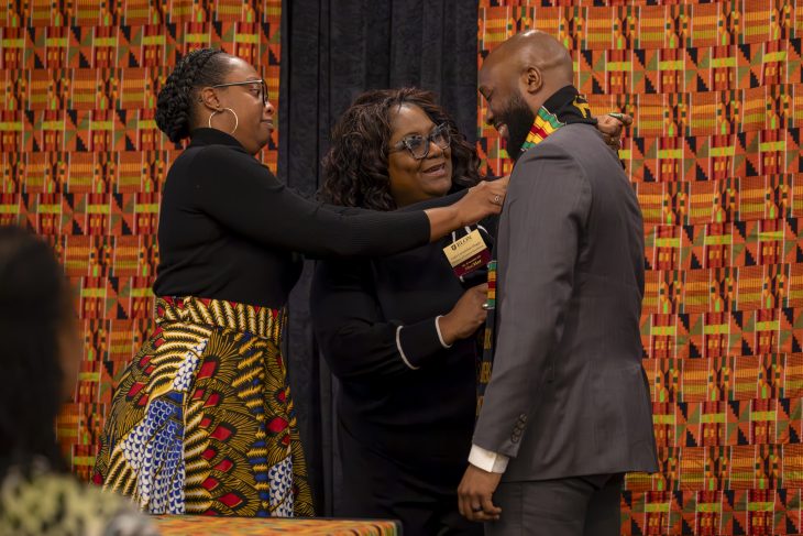 Two women place a kente on a man