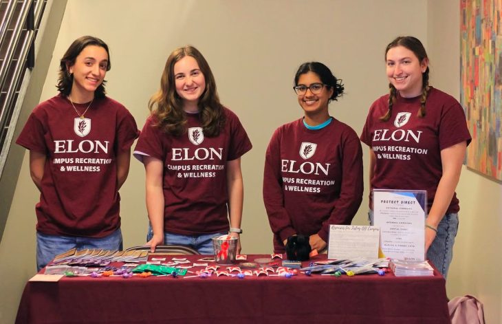 Four students wearing matching maroon Elon Campus Recreation & Wellness shirts stand behind a table filled with informational materials, small giveaways, and a display titled 'Protect Direct.' They are smiling in a brightly lit indoor setting.