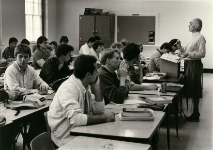 A 1990s classroom at Elon University showing a female professor standing in front of class while students listen from their desks