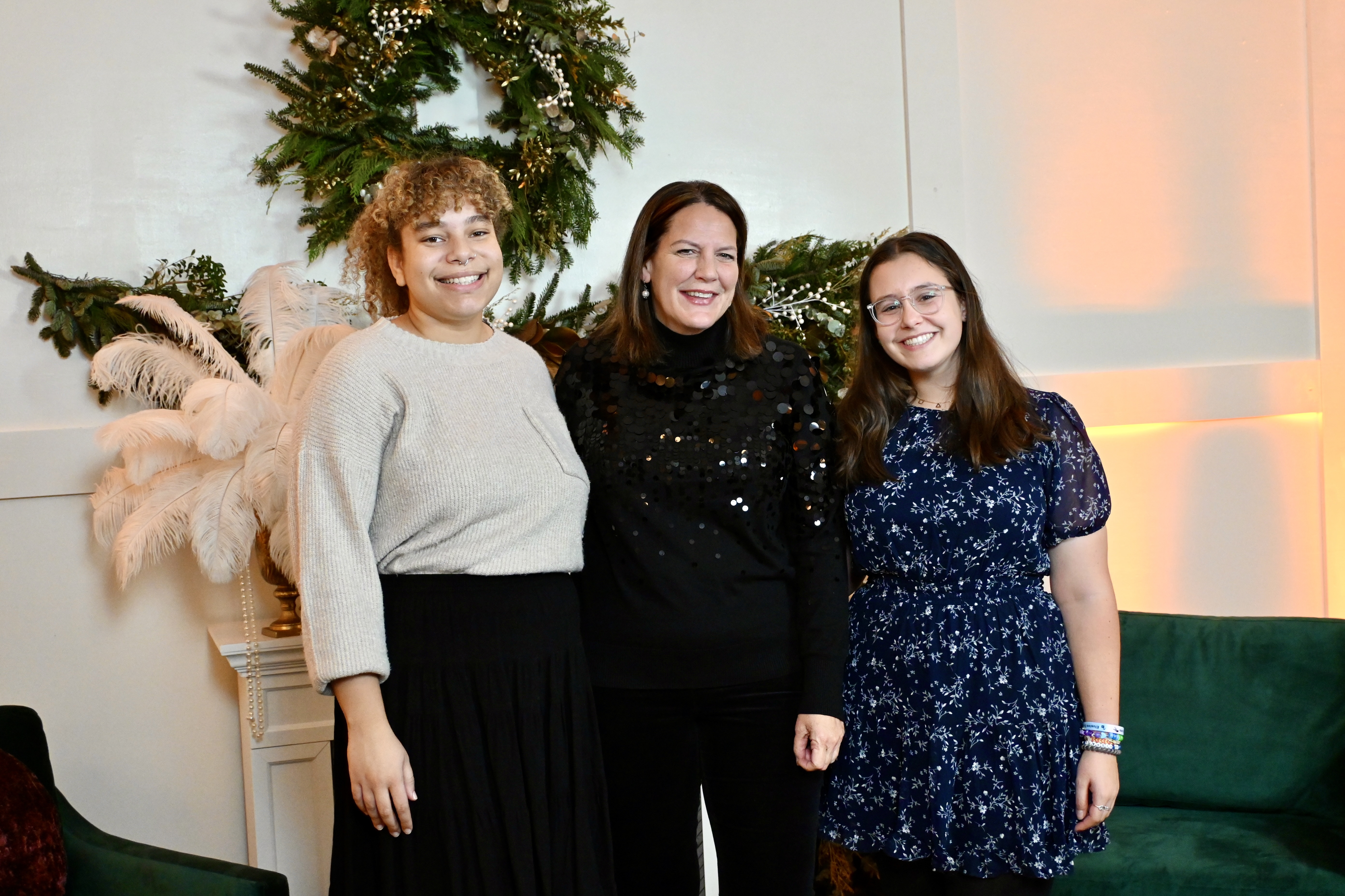 Elon University President Connie Ledoux Book stands smiling between two students at the Student Holiday Party. The group is framed by festive holiday decorations, including a wreath and white feathered accents.