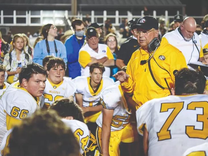 Man in yellow shirt stands and speaks surrounded by football players