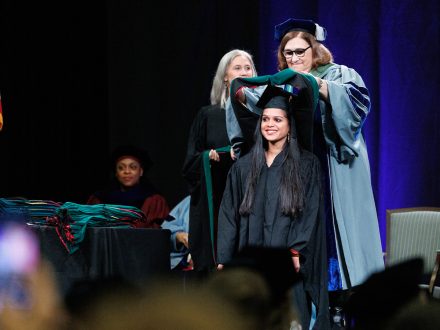 Two women in caps and gowns stand behind a woman in a gap and gown and place a hood over her shoulders.