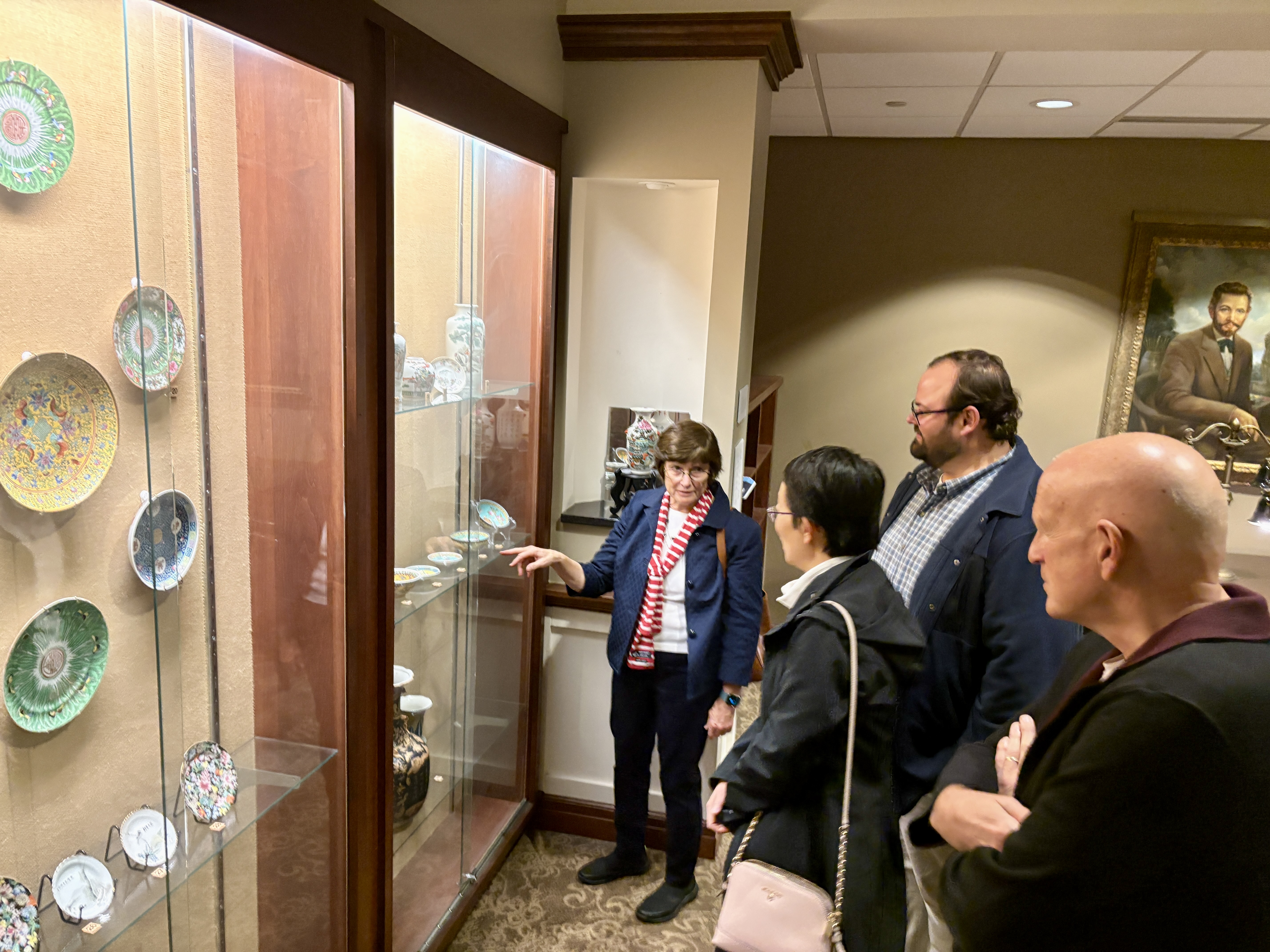 A woman leading a discussion near a display case of decorative plates and vases, pointing towards an artifact as others listen attentively.