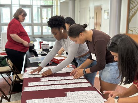 Student volunteers at the 2024 Intersect Conference organize name tags on a maroon tablecloth. Three volunteers focus on arranging the tags, while a staff member in a red shirt stands behind the table. The scene is set in a bright lobby area with large windows.