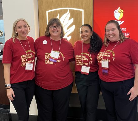 Four staff members from the Center for Leadership pose together at the 2024 Intersect Conference. They are wearing matching red T-shirts with the text "Breaking Boundaries for a Sustainable Future" and conference name tags. They stand in front of a wall featuring a lit emblem and a red digital display.