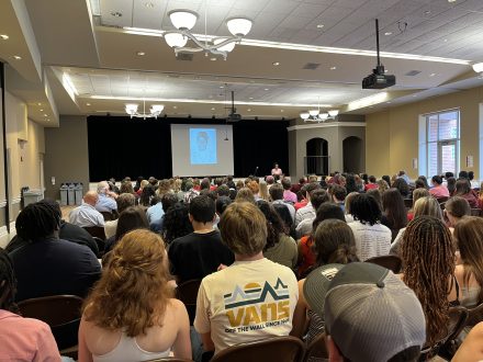 Attendees of the Intersect Conference sit in rows of chairs in a large, well-lit auditorium, listening to the keynote address. A speaker stands at a podium in front of a projection screen displaying a sketch of a person. The audience includes diverse individuals, with some wearing casual attire and others dressed more formally.