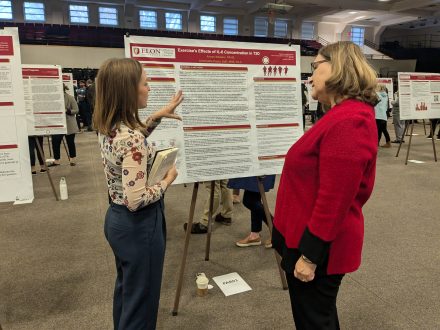 A person stands next to poster board and talks to another person with their hand outstretched