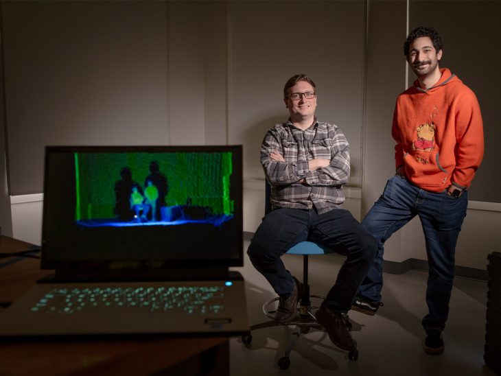 A laptop sits in the foreground showing two human shapes on the screen. In the background is Ryan Mattfield and Rony Dahdal. Mattfield is seated and Dahdal is standing/