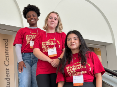 Three student directors of the Intersect Conference stand on a staircase in front of a "Student Centered" sign. From left to right: T,, in black slacks. All wear red conference T-shirts that read “Breaking Boundaries for a Sustainable Future.”