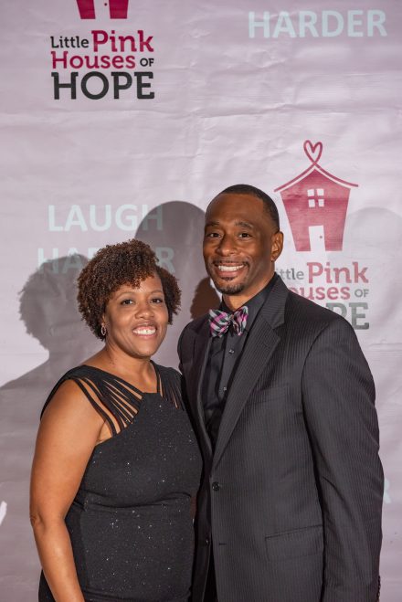 Two people pose for photo in front of pink background that says "Little Pink Houses of Hope"