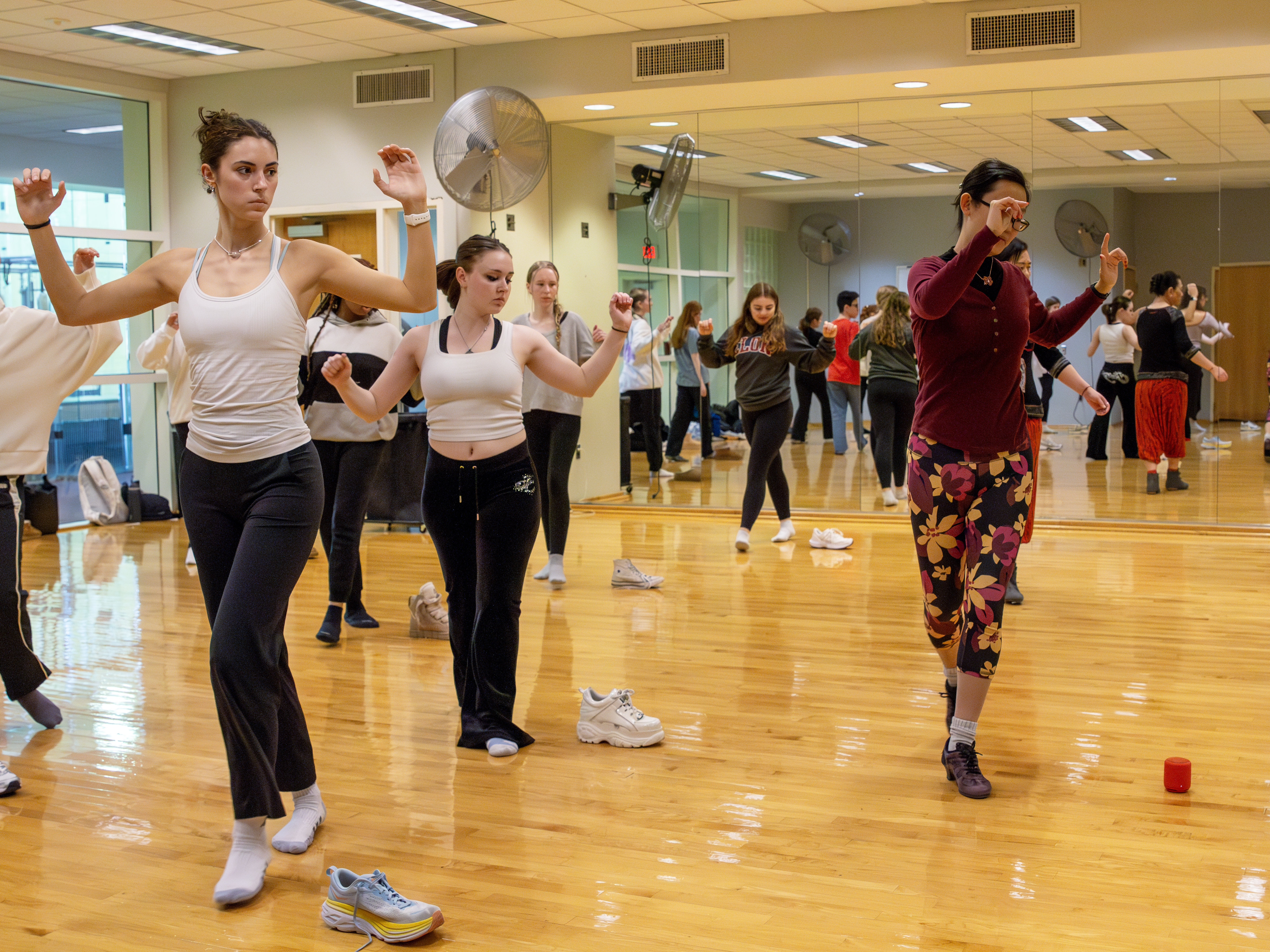 A large group of students in a mirrored dance rehearsal space follow a professor's dance steps