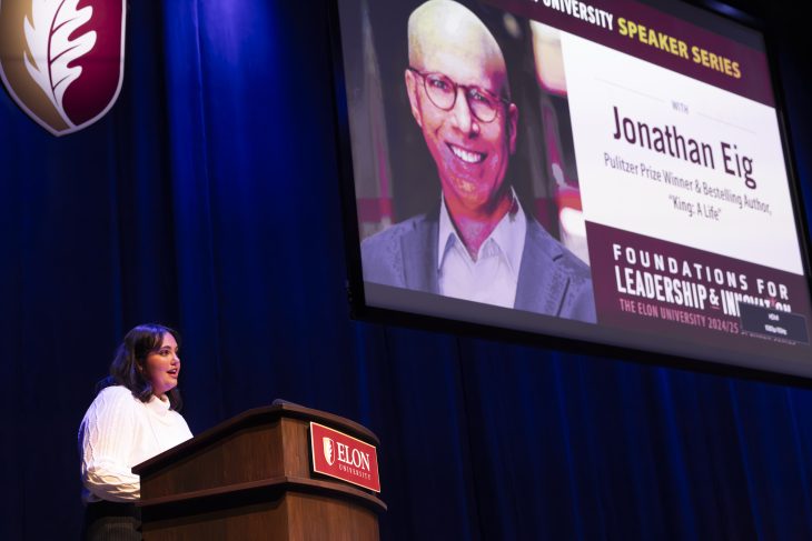 Person speaking at a podium at the Elon University Speaker Series with a large screen behind displaying the name "Jonathan Eig" and a photo of him.