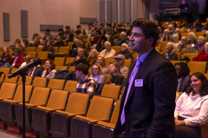 A person wearing a suit with a badge labeled "BRIAN" stands at a microphone before an audience seated in an auditorium.