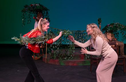 Two actors on stage performing a scene, one holding a branch of greenery extended towards the other amidst a floral set design.